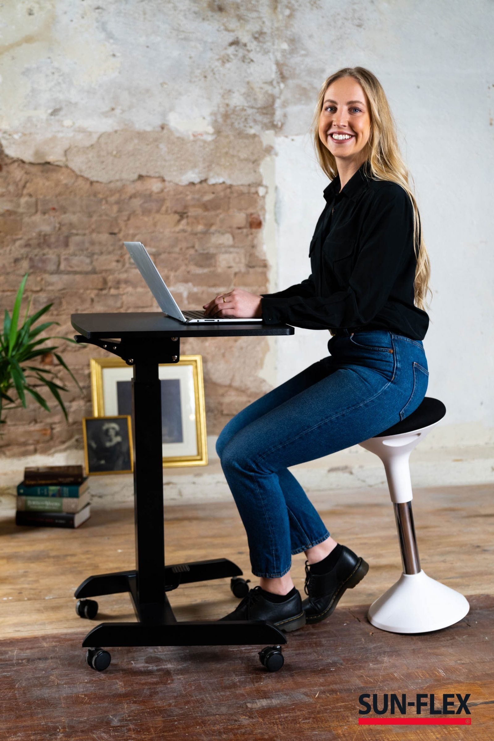 kvinna sitter på  balanspall framför Svart skrivbord med justerbar höjd, idealiskt för begränsade utrymmen och arbete på resande fot.
